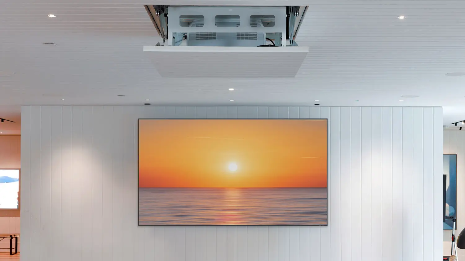 The Carson Phantom Lift Mini installed in a ceiling with a projector mount system partially extended. Below it is a large wall-mounted display showing a sunset over water scene. The room has white paneled walls and recessed lighting.